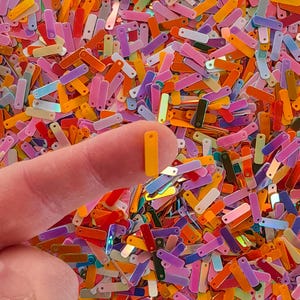 Peut inclure: Gros plan sur une collection de paillettes rectangulaires color&eacute;es dans diverses nuances d'orange, de rose, de violet et de vert. Une paillette jaune est tenue entre un doigt et un pouce. Les paillettes ont une finition brillante et iris&eacute;e.