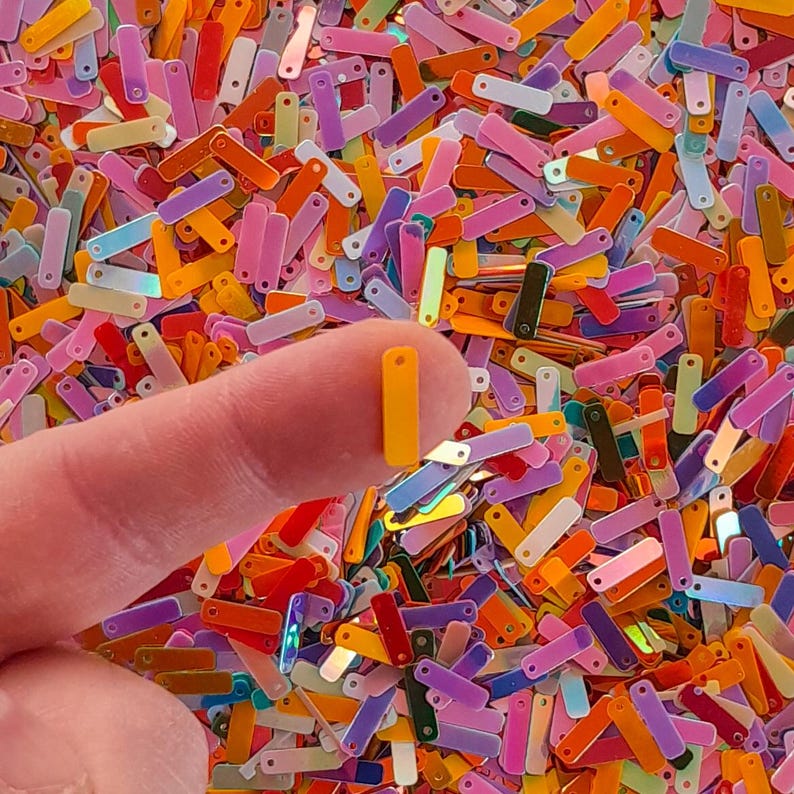 Peut inclure: Gros plan sur une collection de paillettes rectangulaires color&eacute;es dans diverses nuances d'orange, de rose, de violet et de vert. Une paillette jaune est tenue entre un doigt et un pouce. Les paillettes ont une finition brillante et iris&eacute;e.