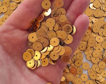 May include: A close-up of a hand holding a pile of gold-colored, round, scalloped sequins. The sequins have a small hole in the center.