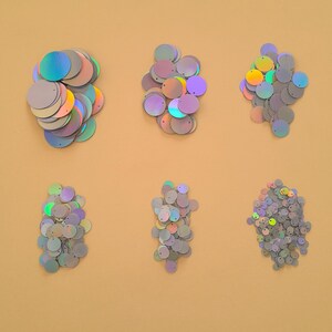 Puede incluir: Surtido de lentejuelas iridiscentes en varios tamaños y formas. Las lentejuelas están dispuestas en seis montones sobre una superficie marrón claro.