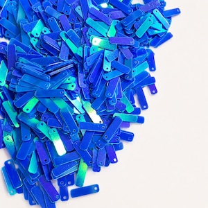 May include: A large collection of small, rectangular, iridescent blue sequins. Each individual sequin features a single circular hole at one end, with a reflective, metallic finish that shifts between shades of deep royal blue and bright cyan.