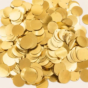 Op de afbeelding: Een grote verzameling platte, ronde goudkleurige metalen schijfjes. Elke schijf heeft een glad, reflecterend oppervlak en een klein gaatje bij de rand, ontworpen voor gebruik als sieraadonderdeel of als materiaal voor knutselprojecten.