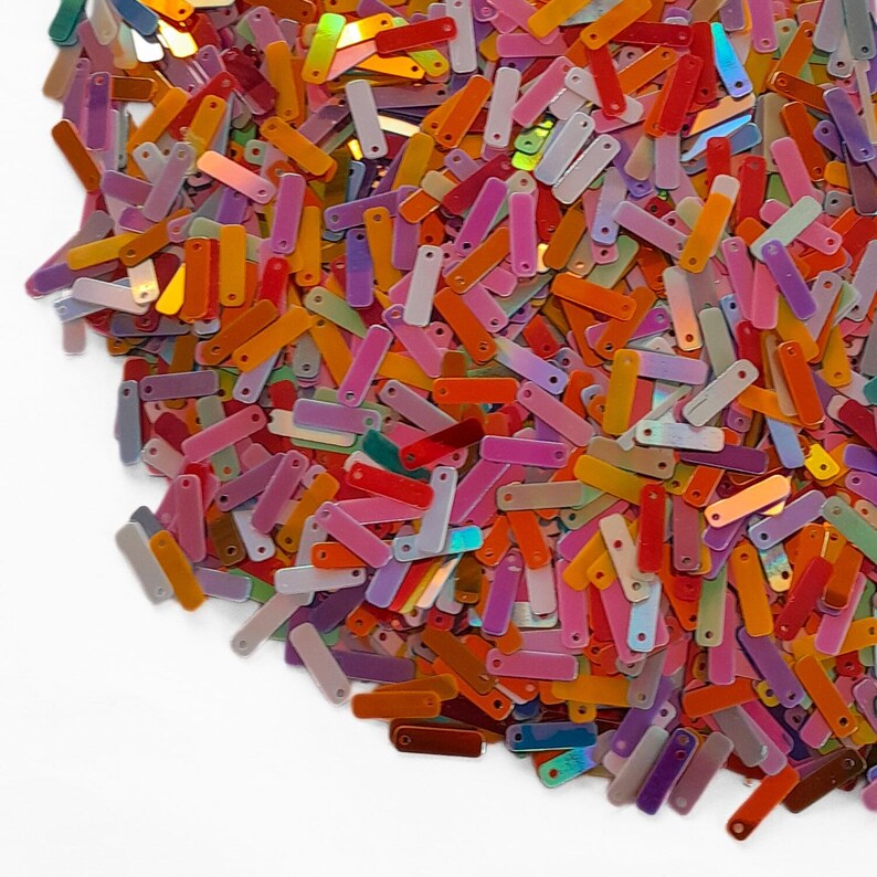 Peut inclure: Gros plan sur une pile de paillettes rectangulaires color&eacute;es. Les paillettes sont dans diverses nuances de rose, orange, jaune, vert et violet. Certaines paillettes ont un &eacute;clat iris&eacute;, refl&eacute;tant la lumi&egrave;re. Les paillettes sont &eacute;parpill&eacute;es sur une surface blanche.