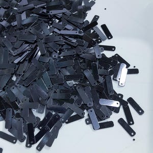 May include: A pile of black rectangular sequins with small holes at the top. The sequins are scattered on a white surface.