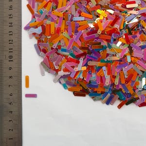 Peut inclure: Une dispersion de paillettes rectangulaires color&eacute;es dans des tons orange, rose, violet et bleu. Une r&egrave;gle avec des graduations en millim&egrave;tres est visible sur le c&ocirc;t&eacute; gauche. Les paillettes sont &eacute;parpill&eacute;es sur une surface blanche.