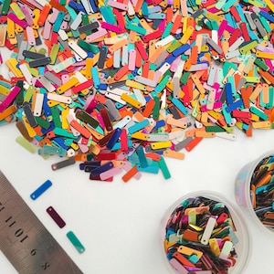 May include: A large pile of colorful, iridescent, rectangular sequins with rounded edges. The sequins are scattered on a white surface and some are in two small clear containers.