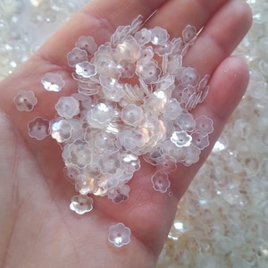 Könnte beinhalten: Ein großer Haufen irisierender weißer, blumenförmiger Pailletten. Die Pailletten sind klein und haben eine leicht erhabene Mitte.