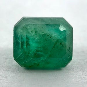 May include: A faceted, emerald-cut emerald gemstone. The gem is a vibrant green color with visible internal inclusions. The cut features a rectangular shape with beveled corners, showcasing the stone's clarity and brilliance. The gemstone is set against a neutral background.