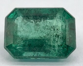 May include: A large, emerald green gemstone with a rectangular, faceted shape. The stone has a cloudy, opaque appearance with visible inclusions.
