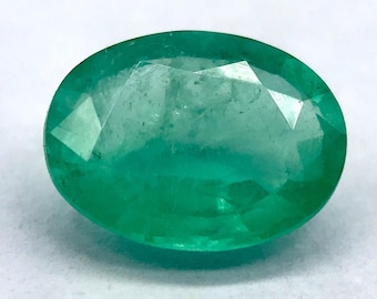 Pode incluir: Uma gema de esmeralda de corte oval, exibindo um tom verde vibrante. A gema tem uma superfície facetada, refletindo a luz e mostrando sua clareza. A esmeralda está sobre um fundo branco, destacando sua cor e forma.