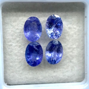 Puede incluir: Cuatro piedras preciosas ovaladas, de color azul-violeta, se muestran en una caja de plástico blanco. Las gemas tienen una superficie facetada, que capta la luz y muestra su color. La caja tiene un interior blanco y una tapa transparente.