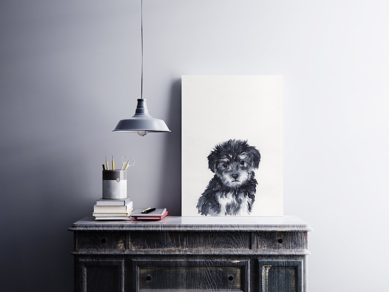 May include: Black and white watercolor painting of a small, fluffy dog sitting on a white background. The painting is framed and hanging on a wall above a dark wooden dresser.
