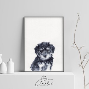 May include: A black and white watercolor painting of a small dog with fluffy fur. The dog is looking at the viewer with a sweet expression. The painting is framed in a black frame and is hanging on a white wall.