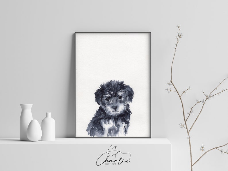 May include: A black and white watercolor painting of a small dog with fluffy fur. The dog is looking at the viewer with a sweet expression. The painting is framed in a black frame and is hanging on a white wall.