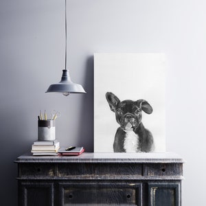 May include: Black and white drawing of a French bulldog on a white canvas, hanging on a wall above a dark wooden dresser with a lamp and books on top.