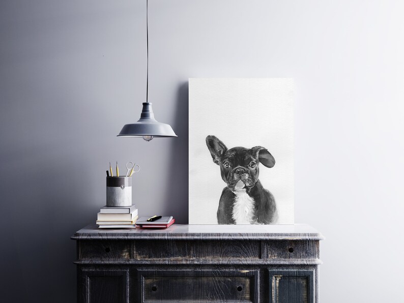 May include: Black and white drawing of a French bulldog on a white canvas, hanging on a wall above a dark wooden dresser with a lamp and books on top.