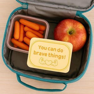 May include: A teal lunchbox containing a clear container of baby carrots, a red apple, and a yellow card with the text "You can do brave things!" and a heart and arm graphic. The lunchbox is open.