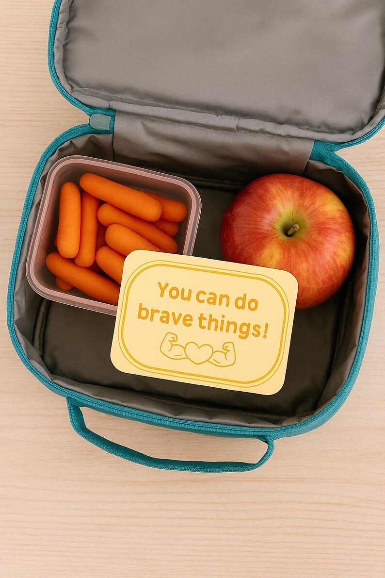 May include: A teal lunchbox containing a clear container of baby carrots, a red apple, and a yellow card with the text "You can do brave things!" and a heart and arm graphic. The lunchbox is open.
