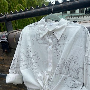 May include: A white long-sleeve button-down shirt featuring a continuous black line art pattern of multiple human faces. The shirt has a classic collar, a front button placket, and is tied at the bottom hem, with sleeves rolled up to the elbows.