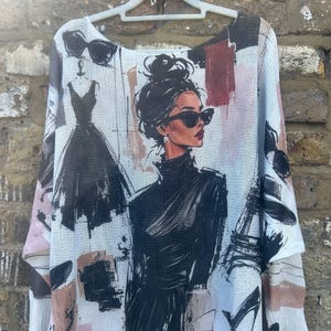 May include: A white long-sleeve jumper featuring a black graphic print of a person with a high bun hairstyle and sunglasses, alongside illustrations of a dress, a perfume bottle, a high-heeled shoe, and abstract pink and dark red brush strokes.