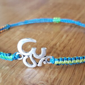 Könnte beinhalten: Ein geflochtenes Armband in Blau, Grün, Orange und Rot mit einem silbernen Anhänger in Form eines Om-Symbols.