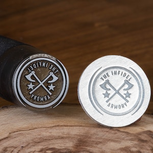 May include: A close-up of two metal stamps with the text "The Infidel Armory" and a crossed axe design. The stamp on the left is a brass-colored metal, and the stamp on the right is a silver-colored metal.