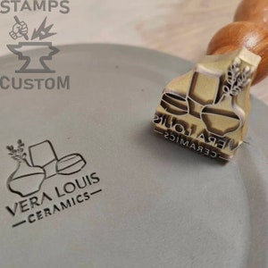 Op de afbeelding: Een messing stempel met de tekst "VERA LOUIS - CERAMICS" en een ontwerp van twee vazen met een takje bloemen. De stempel ligt op een grijs oppervlak.