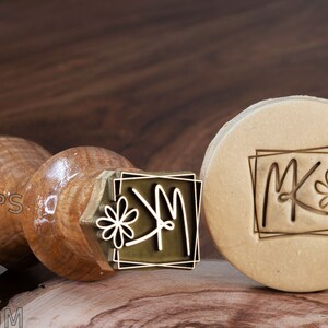 May include: A wooden handle with a brass stamp featuring the letters "MK" with a stylised flower design inside a square frame. The stamp is being used to create an impression on a beige surface.