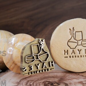 May include: A close-up of two wooden stamps featuring a design of stylised vases and leaves. The stamp on the left is labelled "Hayes Ceramics" and the stamp on the right is labelled "Heyah Ceramics".