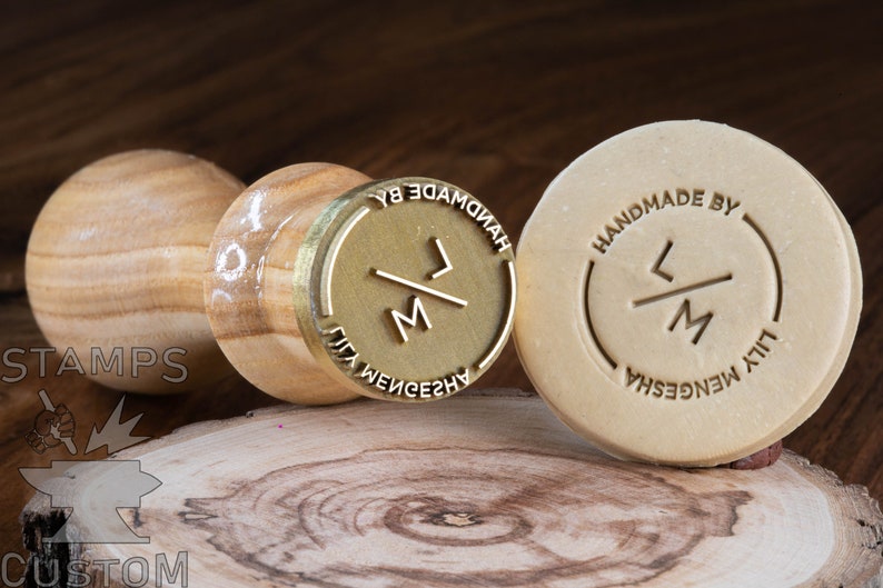 May include: Two wooden stamps with brass inlays. The stamp on the left has a wooden handle and the stamp on the right is a standalone round stamp. Both stamps feature the initials "LM" with a diagonal line through them. The stamp on the left has the text "HANDMADE BY LILY MENGESHA" around the initials. The stamp on the right has the text "HANDMADE BY LILY MENGESHA" around the initials.