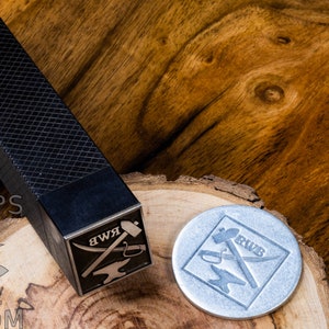 May include: A black metal stamp with a design of a hammer, anvil, and a pair of tongs with the letters RWB. The stamp is sitting on a wooden surface next to a silver metal stamp with the same design.