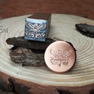 May include: A metal stamp with a design of a black raven with wings spread and the text "BLACK RAVEN" underneath. The stamp is on a wooden surface next to a copper coin with the same design.