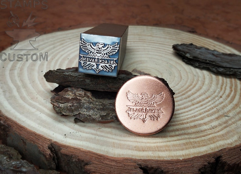 May include: A metal stamp with a design of a black raven with wings spread and the text "BLACK RAVEN" underneath. The stamp is on a wooden surface next to a copper coin with the same design.