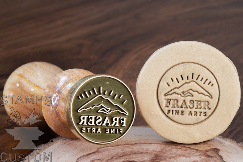 May include: A brass stamp with a wooden handle. The stamp features a mountain range design with a sunburst above it and the text "FRASER FINE ARTS" in a circular design.