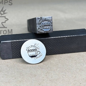 May include: A close-up of two metal stamps. The first stamp is a square with a design of a cup with a handle and the text "Miele zu feiern" and "Studio". The second stamp is a round metal disc with the same design.