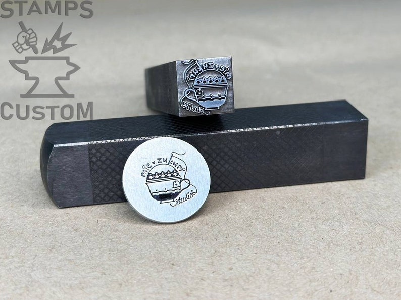 May include: A close-up of two metal stamps. The first stamp is a square with a design of a cup with a handle and the text "Miele zu feiern" and "Studio". The second stamp is a round metal disc with the same design.