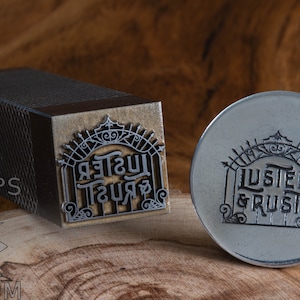 May include: A metal stamp with the words "LUSTER & RUST" in a decorative font. The stamp is in the shape of a gate with a crown on top. The stamp is on a wooden surface.