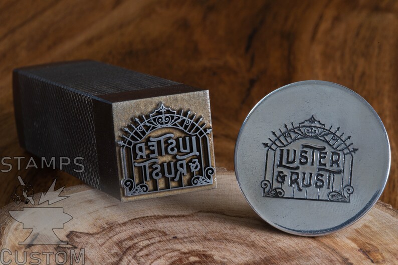 May include: A metal stamp with the words "LUSTER & RUST" in a decorative font. The stamp is in the shape of a gate with a crown on top. The stamp is on a wooden surface.