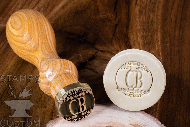 May include: A wooden handle attached to a brass stamp with the initials "CB" and the words "Handmade Pottery" in a wreath design. The stamp is used to create an impression on a piece of clay.