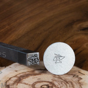 May include: A metal stamp with a design of a blacksmith's anvil and tools. The stamp is on a wooden surface. The stamp is labeled "A.R."