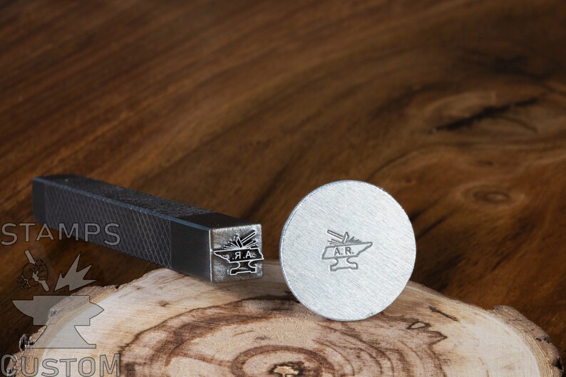 May include: A metal stamp with a design of a blacksmith's anvil and tools. The stamp is on a wooden surface. The stamp is labeled "A.R."