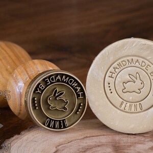 May include: A wooden handle attached to a brass stamp with a circular design. The design features a rabbit silhouette and the text "HANDMADE BY HENNO". The stamp is used to imprint the design onto a bar of soap.