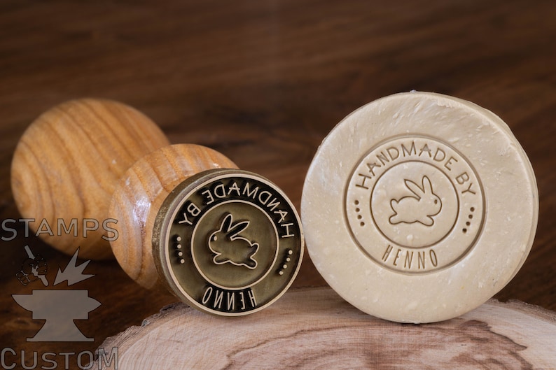 May include: A wooden handle attached to a brass stamp with a circular design. The design features a rabbit silhouette and the text "HANDMADE BY HENNO". The stamp is used to imprint the design onto a bar of soap.
