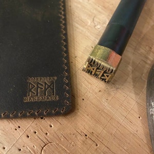 May include: A close-up of a leather journal with a stitched edge and the text "AMERICAN RFM HANDMADE" embossed on the cover. A brass stamp with the text "AMERICAN RFM HANDMADE" is also shown.