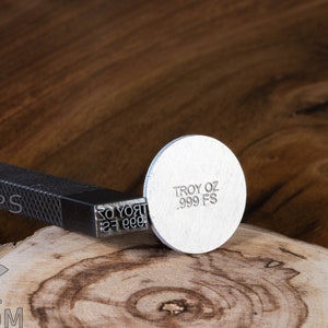 May include: A silver metal stamp with the text "TROY OZ .999 FS" on a wooden surface.