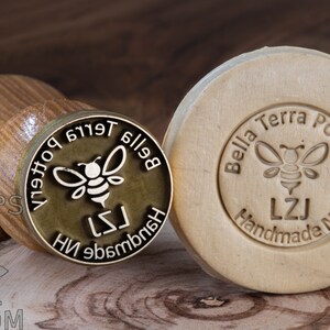May include: Two round, wooden stamps featuring a bee design. The stamp on the left is brass with the text "Bella Terra Pottery" in reverse. The stamp on the right is made of wood with the text "Bella Terra Pottery, LZJ Handmade NH" in a circular design.