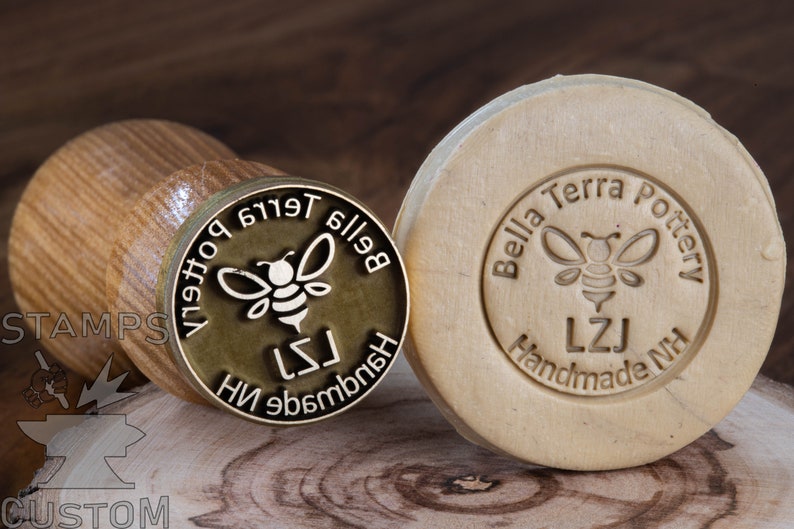 May include: Two round, wooden stamps featuring a bee design. The stamp on the left is brass with the text "Bella Terra Pottery" in reverse. The stamp on the right is made of wood with the text "Bella Terra Pottery, LZJ Handmade NH" in a circular design.