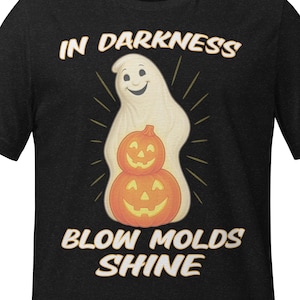 Puede incluir: Camiseta negra con un diseño de Halloween. El diseño presenta un fantasma sonriente sobre dos calabazas apiladas, con el texto "IN DARKNESS BLOW MOLDS SHINE" en una fuente vintage. La camiseta está hecha de un material suave.