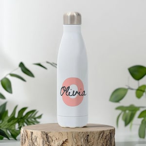 以下が含まれることがあります： シルバーのキャップが付いた白い断熱ウォーターボトル。ボトルには、コーラル色の円の上に黒い筆記体で「Olivia」という名前が書かれています。ボトルは木の台の上に置かれ、背景には緑の葉があります。ボトルの高さは約25cmです。