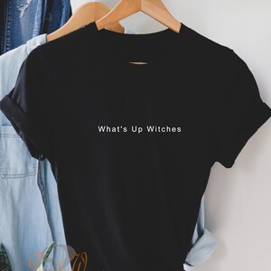 May include: A black short-sleeved t-shirt with a crew neckline and rolled sleeves. The centre of the chest features the white text "What's Up Witches" printed in a simple, clean sans-serif font. The garment is displayed on a wooden hanger.